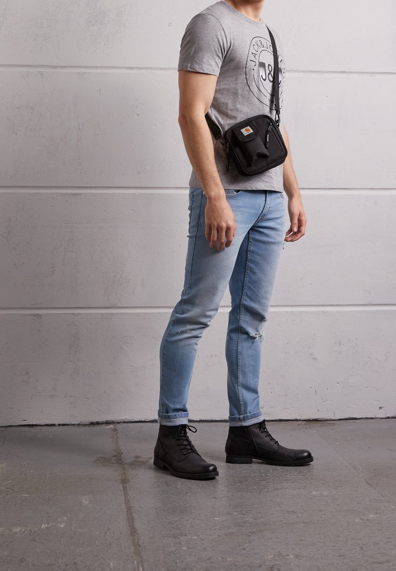 Bolso cruzado negro con cremalleras, llevado sobre una camiseta gris y unos jeans desgastados de color azul claro, combinado con botas negras de cordones.