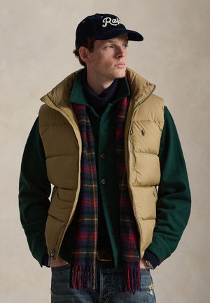 Jeune homme portant une veste matelassée beige, une veste verte, un pull bleu marine, une écharpe à carreaux, un jean bleu et une casquette de baseball bleu marine, regardant sur le côté les mains dans les poches.