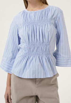 Vrouw draagt een lichtblauwe, witgestreepte blouse met geplooide smock en brede manchetten, gecombineerd met beige broek.