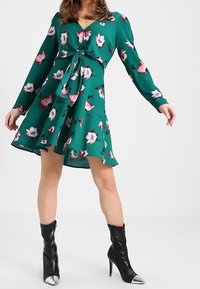 Žena v zelených květinových šatech s dlouhými rukávy a předním uzlem, doplněná černými vysokými botami na podpatku se stříbrnými špičkami.