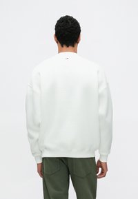 Bílá mikina s kulatým výstřihem a spadlými rameny. Měkký materiál a žebrované manžety. Malý detail loga na zadním límci. Kombinováno s olivově zelenými kalhotami.