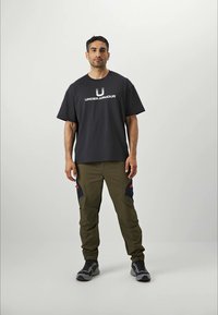 Homme se tenant de face, portant un t-shirt noir Under Armour, un pantalon cargo olive avec des patchs noirs et des baskets grises sur un fond uni.
