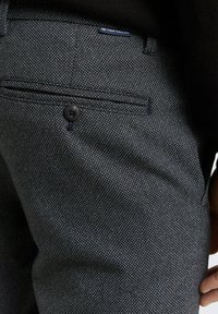 Dark grey textured trousers with a subtle pattern, featuring a button closure and a small branded tag. The fabric appears durable and structured.