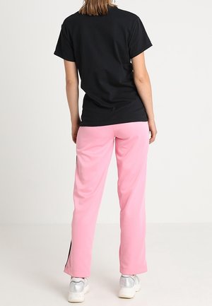 Personne portant un t-shirt noir à manches courtes, un pantalon rose avec des bandes latérales noires et des baskets blanches, debout devant un fond blanc uni.