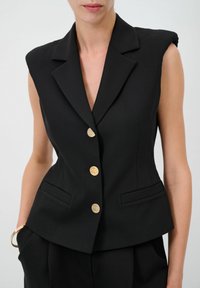 Schwarzer ärmelloser Blazer mit tiefem V-Ausschnitt, goldfarbenen Knöpfen und zwei Fronttaschen. Taillierte Passform mit strukturierten Schultern und glattem Stoff.