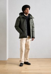 Parka verde esponjosa con capucha, detalles en negro y múltiples bolsillos. Combinada con pantalones beige y zapatos negros sin cordones. Fondo de suelo de madera.