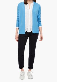 Modrý kardigan vyrobený z lehkého materiálu, vrstvený přes bílou blůzu s vyšíváním, kombinovaný s černými kalhotami a bílými teniskami.