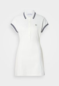 Robe polo blanche à manches courtes, col rayé bleu marine, patte de boutonnage à quatre boutons et logo crocodile vert brodé sur la poitrine gauche.