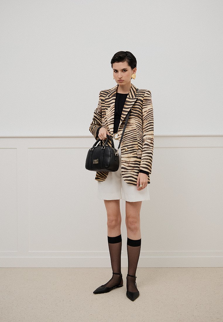 Femme portant un blazer à rayures de tigre, un top noir, un short blanc, des chaussettes transparentes montantes et des ballerines noires, tout en tenant un sac à main noir.