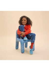 Niño sentado en una silla azul moteada, vistiendo un suéter naranja, zapatos azules y jeans de mezclilla enrollados con calcetines rojos, sonriendo mientras abraza sus rodillas.