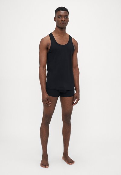 Schwarzes Baumwoll-Tanktop mit breiten Trägern, kombiniert mit schwarzen enganliegenden Unterhosen. Das Material wirkt glatt und hat ein minimalistisches Design.