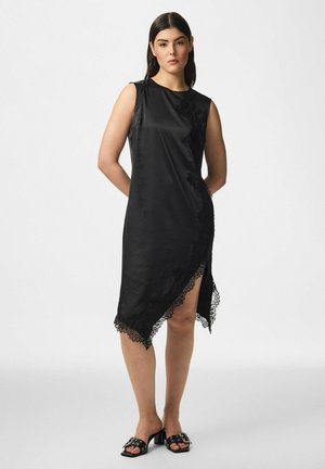 Un mannequin portant une robe noire sans manches, à l’ourlet asymétrique bordé de dentelle, et des sandales noires cloutées, se tient devant un fond de studio blanc.