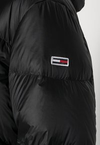 Zwarte pufferjack met gewatteerde textuur, voorzien van een opvallend geborduurd logo in het wit, rood en blauw op de linkermouw.