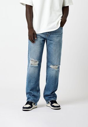 Jeans rotos de color azul claro con un corte holgado, que cuentan con parches deshilachados en las rodillas. Combinados con zapatillas negras y blancas y una camiseta blanca holgada.