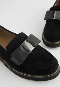 Mustat mokkanahka loaferit, joissa on näkyvä nahkavekki ja metallinen yksityiskohta. Kenkäparissa on pehmustettu pohja ja kumipohja, jossa on erottuva ommel.