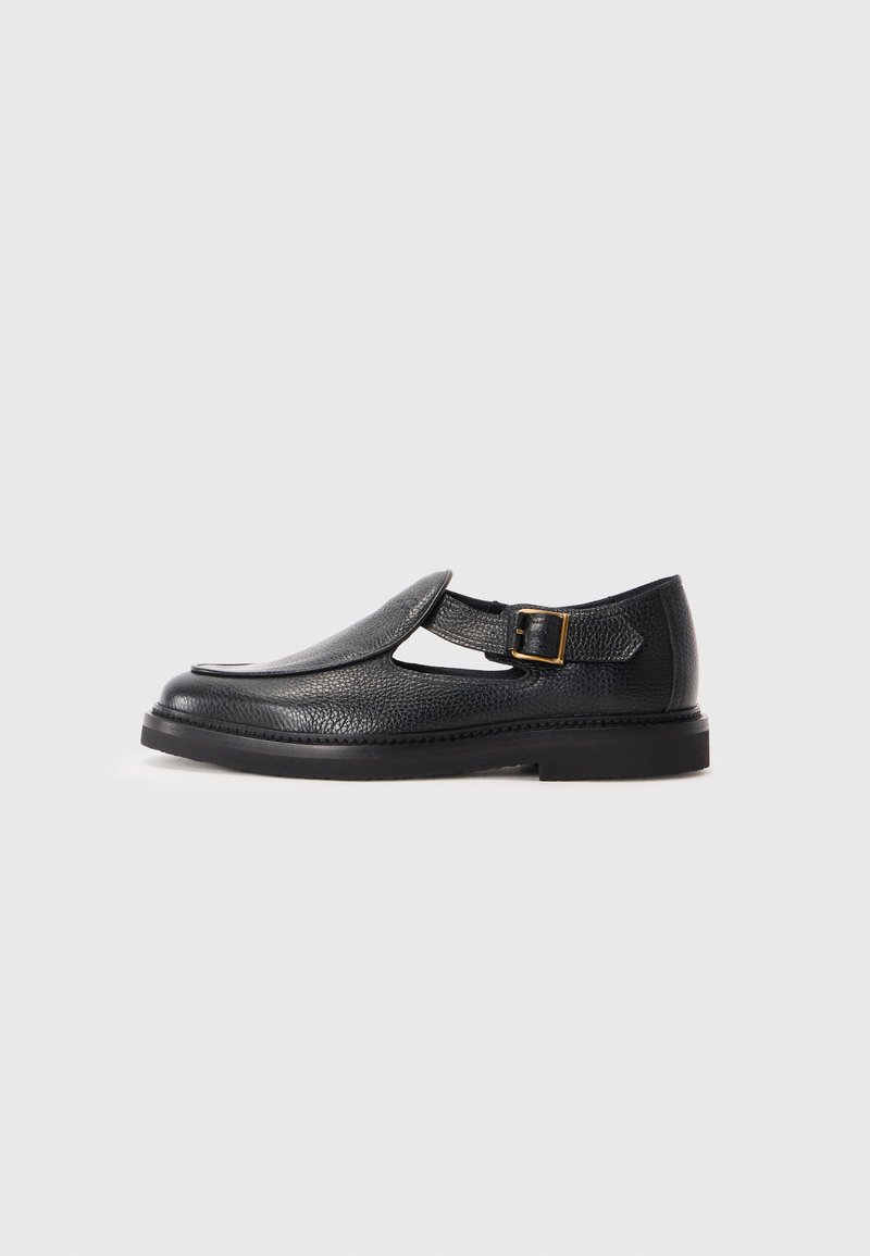 Chaussure T-bar en cuir noir avec une finition texturée, dotée d'une sangle à boucle et d'une pointe arrondie. Semelle noire basse et robuste.
