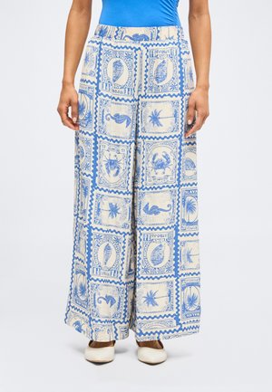 Pantalon à jambes larges avec motifs bleus de timbres-poste vintage représentant des créatures marines et des palmiers, porté avec des chaussures blanches et un haut bleu.