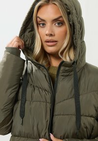 Veste matelassée vert olive avec capuche, dotée d'une fermeture éclair, de cordons de serrage noirs et d'un motif matelassé. Léger, texture douce.