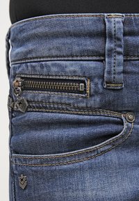 Džínové kalhoty v tmavě modré barvě s předním kapsou na zip, stříbrnými kovovými prvky, kontrastním prošíváním a mírně opraným vzhledem.