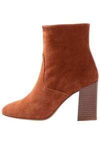 Botte de cheville en daim brun avec un bout pointu, un haut ajusté et un talon en bloc empilé avec des rainures horizontales. Texture lisse.