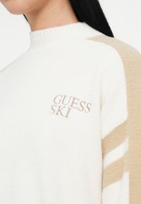 Fehér pulóver bordás gallérral, bézs díszítéssel és világos színben hímzett "GUESS SKI" felirattal. Puha tapintás és laza fazon.