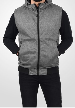 Homme portant un gilet sans manches à capuche gris zippé sur un t-shirt noir à manches longues, mains dans les poches, sur fond blanc.