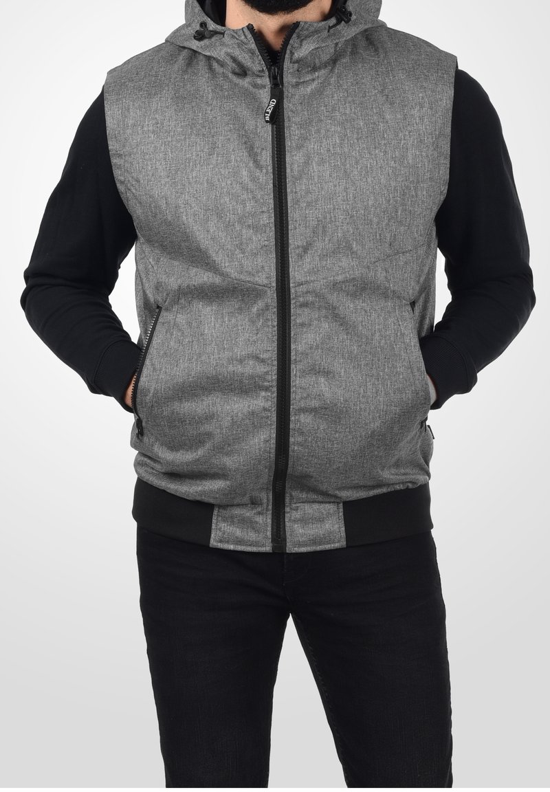 Homme portant un gilet sans manches à capuche gris zippé sur un t-shirt noir à manches longues, mains dans les poches, sur fond blanc.