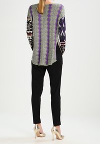 Maglia a maniche lunghe con un corpo verde e motivi zigzag viola, maniche nere a fantasia; abbinata a pantaloni neri e tacchi dorati.