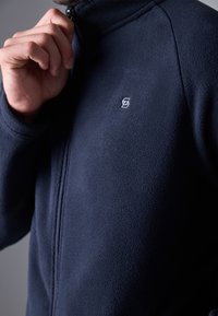 Jūras spēku flīsa jaka ar augstu apkakli, pilnu rāvējslēdzēju un izšuvu logotipu uz krūtīm. Mīksta tekstūra ar vienmērīgu apdari.