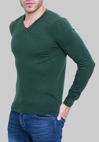 Pull à col en V vert en tissu doux, coupe ajustée, manches longues et poignets côtelés. Associé à un jean en denim bleu.
