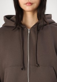 Sudadera con cremallera marrón, con un bolsillo frontal, capucha con cordón y detalles de cremallera plateados, confeccionada en una tela texturizada.