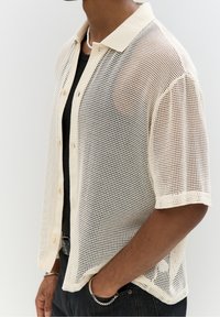 Mężczyzna w beżowej koszuli z krótkim rękawem, zapinanej na guziki, nosi czarną bluzkę, czarne dżinsy oraz srebrną bransoletkę łańcuszkową, a rękę trzyma w kieszeni.