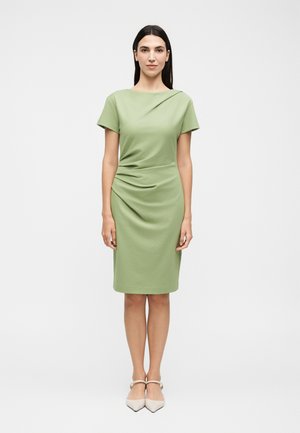 Γυναίκα που στέκεται αντιμετωπίζοντας μπροστά, με ανοιχτοπράσινο φόρεμα μέχρι το γόνατο με κοντά μανίκια και λευκά μυτερά παπούτσια.