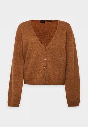 Une femme porte un cardigan marron sur un chemisier crème à nœud et un pantalon à jambes larges. Elle tient un sac à main marron devant un fond de studio uni.