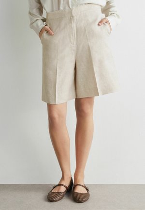 Personne portant un short beige sur mesure jusqu'aux genoux, une blouse blanche, les mains dans les poches, et des chaussures Mary Jane tressées marron.