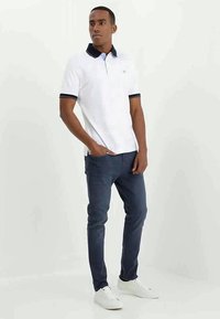 Camisa polo branca com colarinho e punhos em azul marinho, combinada com jeans azul-escuro e ténis brancos. O modelo está em um fundo neutro.
