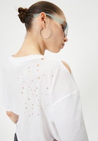 Hvid bomuld T-shirt med slidte huller i forskellige størrelser på ryggen. Har et afslappet fit, rullede ærmer og en let tekstur.