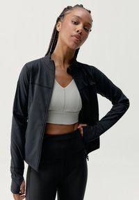 Czarna sportowa kurtka z zamkiem, zaprojektowana w dopasowanym kroju, zestawiona z jasnoszarym stanikiem sportowym i czarnymi legginsami z wysokim stanem.