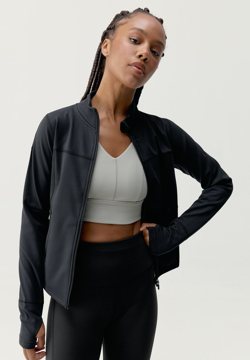 Czarna sportowa kurtka z zamkiem, zaprojektowana w dopasowanym kroju, zestawiona z jasnoszarym stanikiem sportowym i czarnymi legginsami z wysokim stanem.