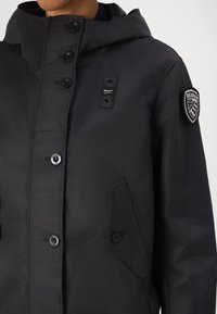 Veste imperméable noire avec capuche, dotée d'un zip à l'avant, de plusieurs boutons, d'un logo sur la poitrine et d'une poche latérale inclinée. Texture lisse.