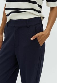 Navy trousers with a smooth texture, featuring front pleats and side pockets. Paired with a striped knitted top.