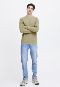 Pulover din tricot ribat de culoare verde măsliniu, combinat cu blugi drepti de culoare albastru deschis și adidași bej. Modelul stă cu brațele încrucișate pe un fundal alb.