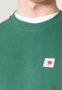 Sudadera verde con cuello redondo de canalé, con un cuello interior blanco y un pequeño parche cuadrado con un símbolo rosa en el pecho.