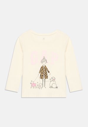Chemise crème à manches longues avec une femme dessinée en manteau léopard marchant avec trois chiens, dont un portant un pull rose, avec de faibles lettres GAP au-dessus.
