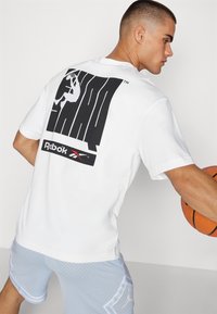 Hvit bomulls t-skjorte med et stort svart grafikk og Reebok-logo på baksiden, paired med lyseblå perforerte shorts.