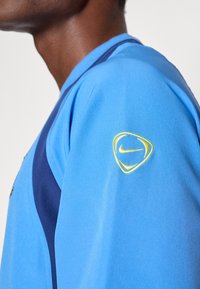 Zils sporta krekls ar tumši zili akcentiem un gludu tekstūru. Uz kreisā pleca ir dzeltenais Nike logo.