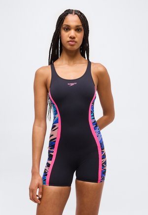 Moteris su pintais plaukais, dėvinti juodą Speedo maudymosi kostiumėlį su rožinėmis juostelėmis ir mėlynos spalvos raštuotais šonais, stovinti prieš vientisą foną.