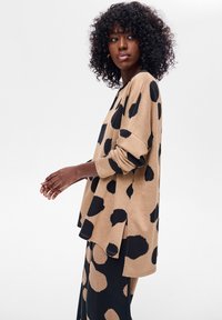 Tanowy, oversize'owy sweter z dużymi czarnymi plamami, z długimi rękawami i bocznymi rozcięciami. Zestawiony z czarnym wzorzystym dołem.