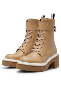 Botas de tobillo de cuero beige con cordones en la parte frontal, punta redonda, suela gruesa blanca y una correa lateral con detalle de hardware metálico.