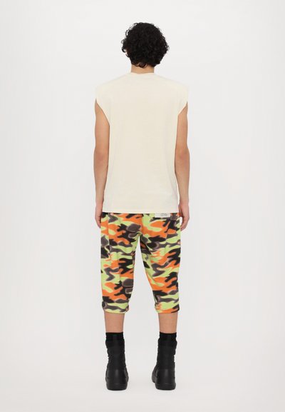 Beige ermeløs skjorte, camo-mønstrede sweatpants i grøn, orange og sort, kombinert med svarte ankelstøvler. Vist bakfra.